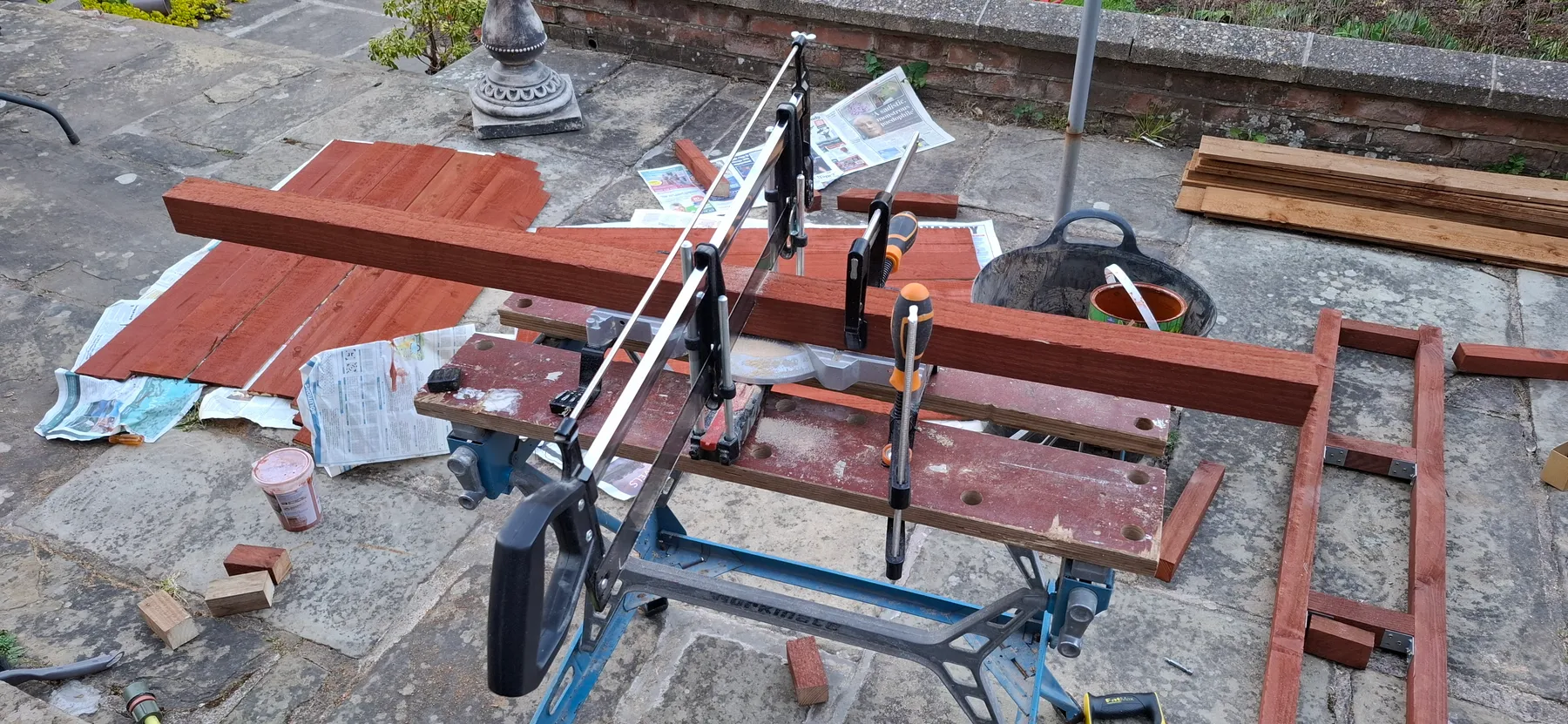 Timber lengths clamped to a mitre saw stand, mid-cut, with stain drying on cladding boards nearby